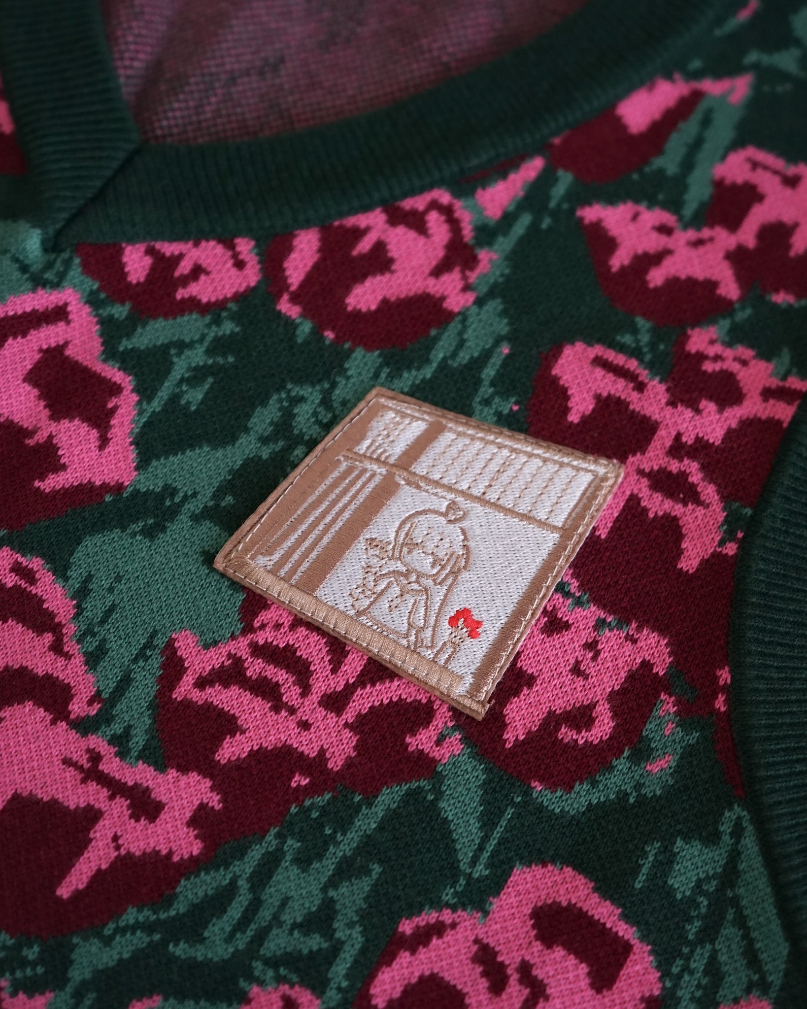 a closeup shot of a green sweater with pink rose floral pattern and an embroidered patch featuring an anime girl standing by a window with a red flower in her hand.