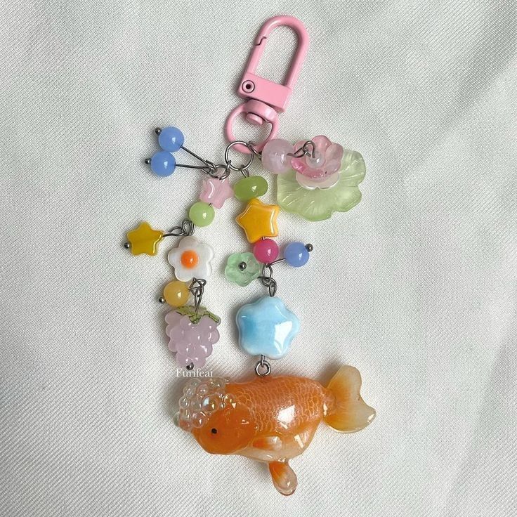 a charm made of beads hangs from a pink clasp. the charm includes beads of different colors and shapes, including stars, flowers, and fruit. at the bottom of the charm is an orange fish.