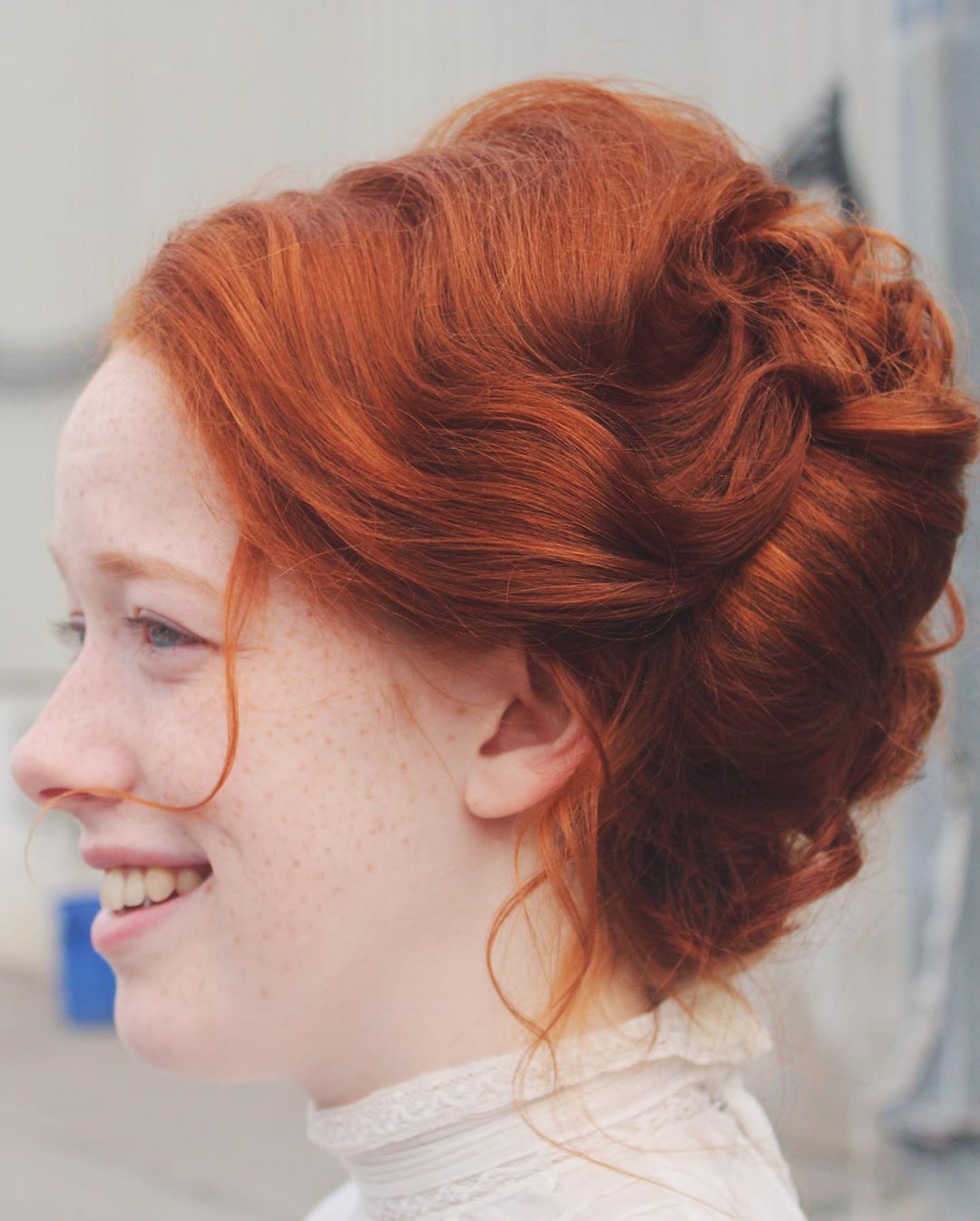 the image is a closeup profile shot of a young woman with vibrant, curly, reddishorange hair styled in a loose, elegant updo.  her hair is voluminous and features soft waves and curls. she has pale skin with visible freckles and is wearing a white, lacy collared top. the background is blurred and out of focus, drawing attention to the subject. the overall aesthetic is romantic and slightly vintage, suggesting a period hairstyle.