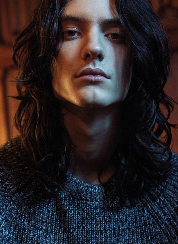 a portrait of a man with long dark hair wearing a gray sweater. the lighting highlights his strong jawline and serious expression.