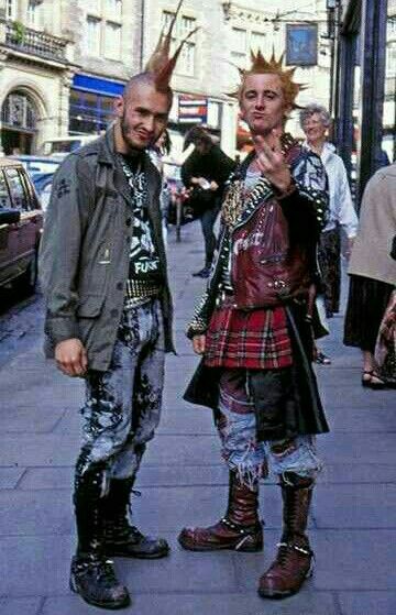 two punks with mohawks stand on a city street. one is wearing a khaki jacket, a tshirt with a "punk" graphic, and patterned jeans with black combat boots. the other is wearing a studded leather jacket over a tartan skirt and distressed jeans with brown combat boots. they are both in punk fashion with distinct hairstyles and clothing choices. the background shows a city street with buildings and other people.
