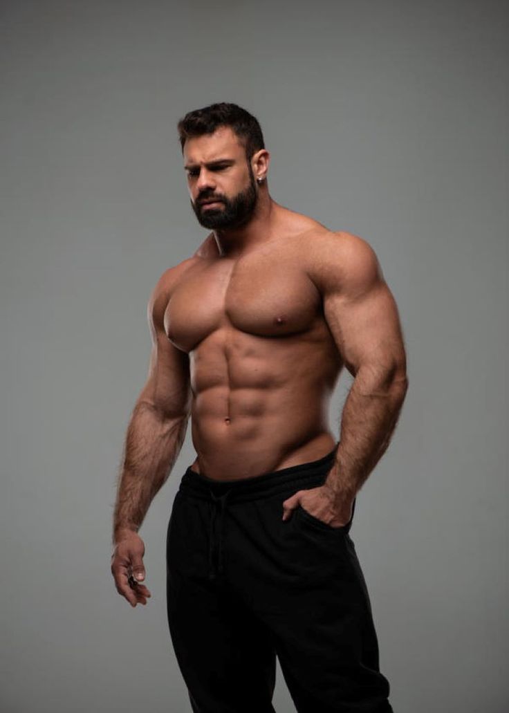 a muscular man is standing with his arms at his side. he is shirtless and wearing black sweatpants. he has a six pack and a muscular chest. he is looking down at the floor. he is standing in front of a grey background.  the man is handsome and attractive.