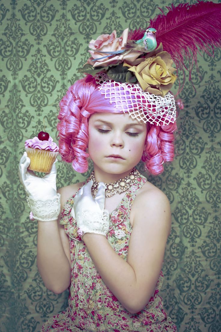 a young child dressed in a vintage fashion style, wearing a pink wig, a hat decorated with flowers, a feather, and a bird. she is holding a cupcake in one hand and wearing white gloves, a floral dress, and a necklace. she has makeup on and a mole on her face, and her eyes are closed. the background is a green patterned wallpaper.