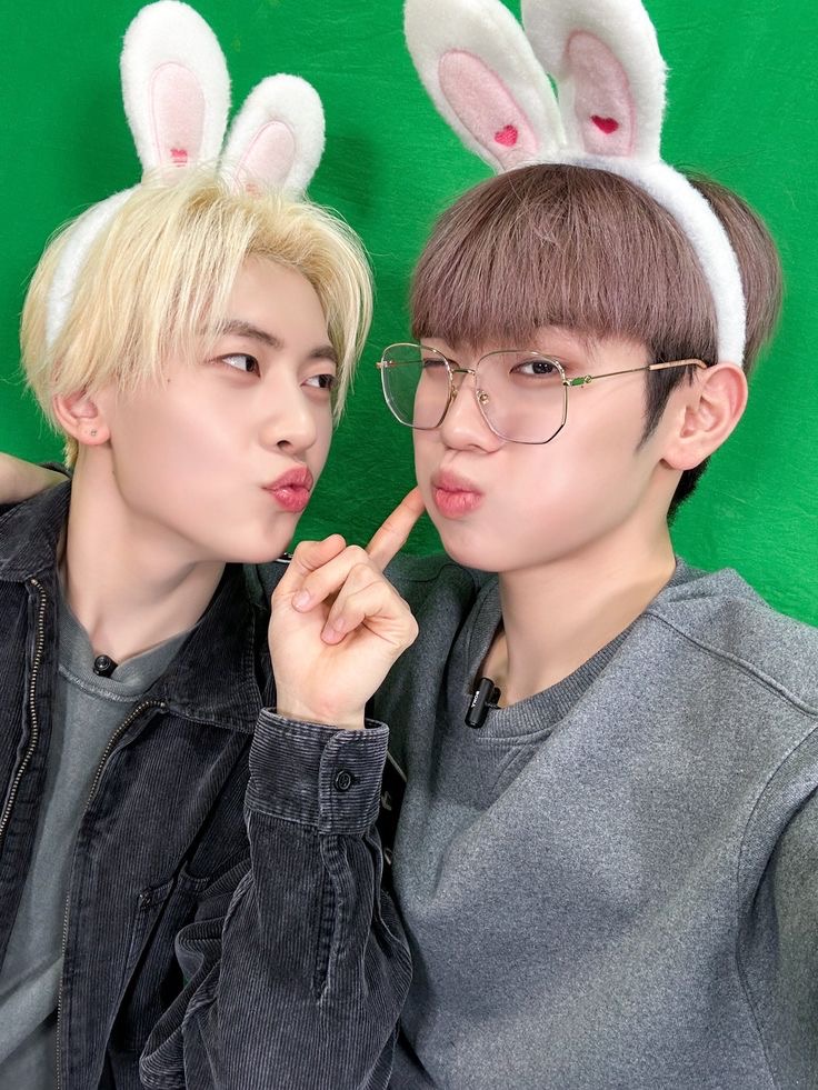 two young asian men pose playfully for a selfie in a studio. they are wearing matching bunny ear headbands and making pouting expressions. the man on the left has blond hair and a black jacket, while the man on the right has brown hair and a gray sweatshirt. the background is a bright green color, typical of a studio setting. the lighting is soft and artificial.