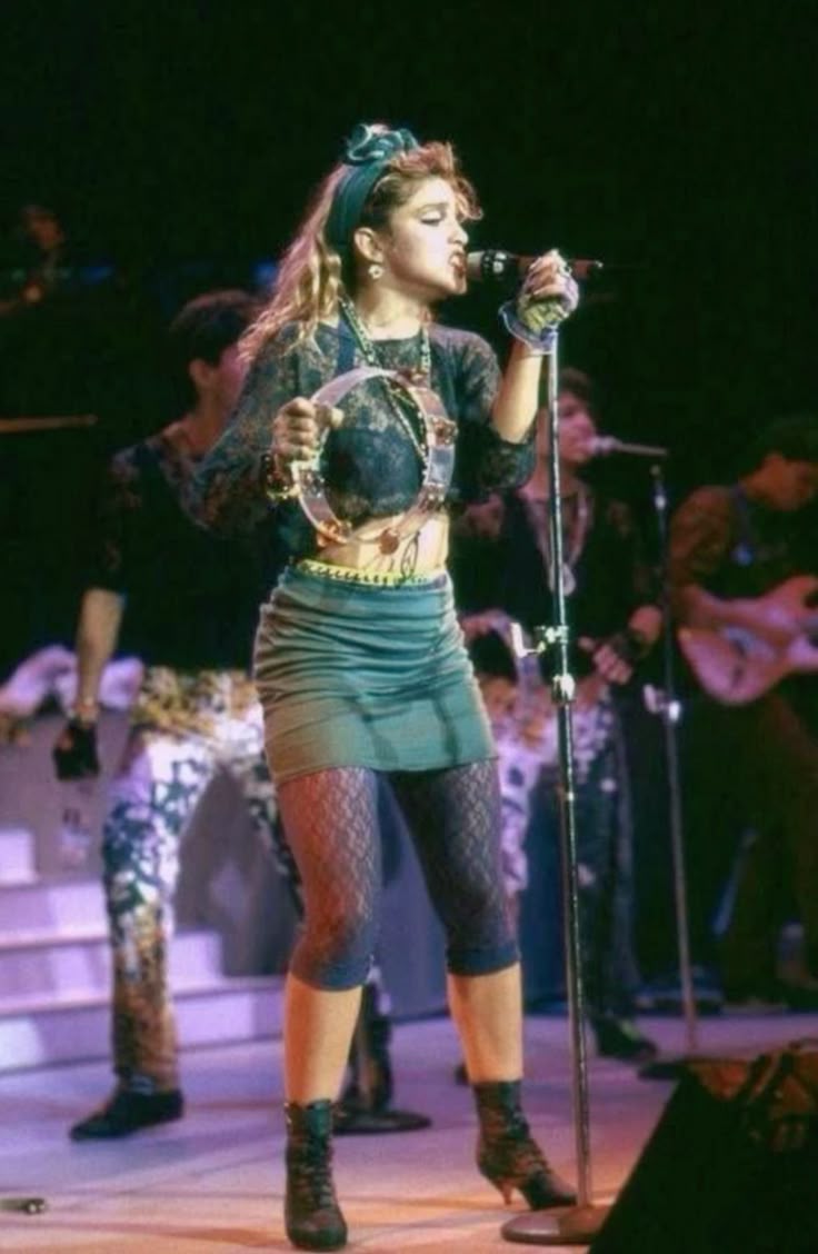 the image shows madonna performing on stage. she is holding a tambourine in her left hand and a microphone in her right hand. she is wearing a dark lace top, a green miniskirt, lace leggings and black boots. she also has a green headband. there are other musicians in the background.