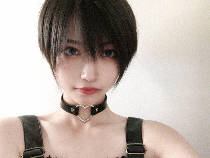 a portrait of a young asian woman with short black hair, wearing a heartshaped choker and a black strapped top. she's looking directly at the camera.  the background is a plain white wall.  the woman has fair skin and a  sweet expression.  the lighting is soft, and the photo is wellcomposed.