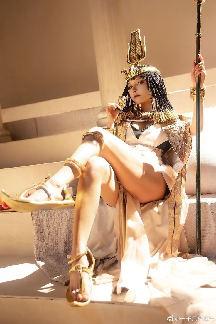 a woman dressed in a cleopatra cosplay sits on a throne. she wears a headdress with an elaborate gold design and is holding a staff. she wears gold jewelry, including bracelets, arm cuffs, and anklets. she is also wearing gold sandals. the throne is white and the background is a neutral color.