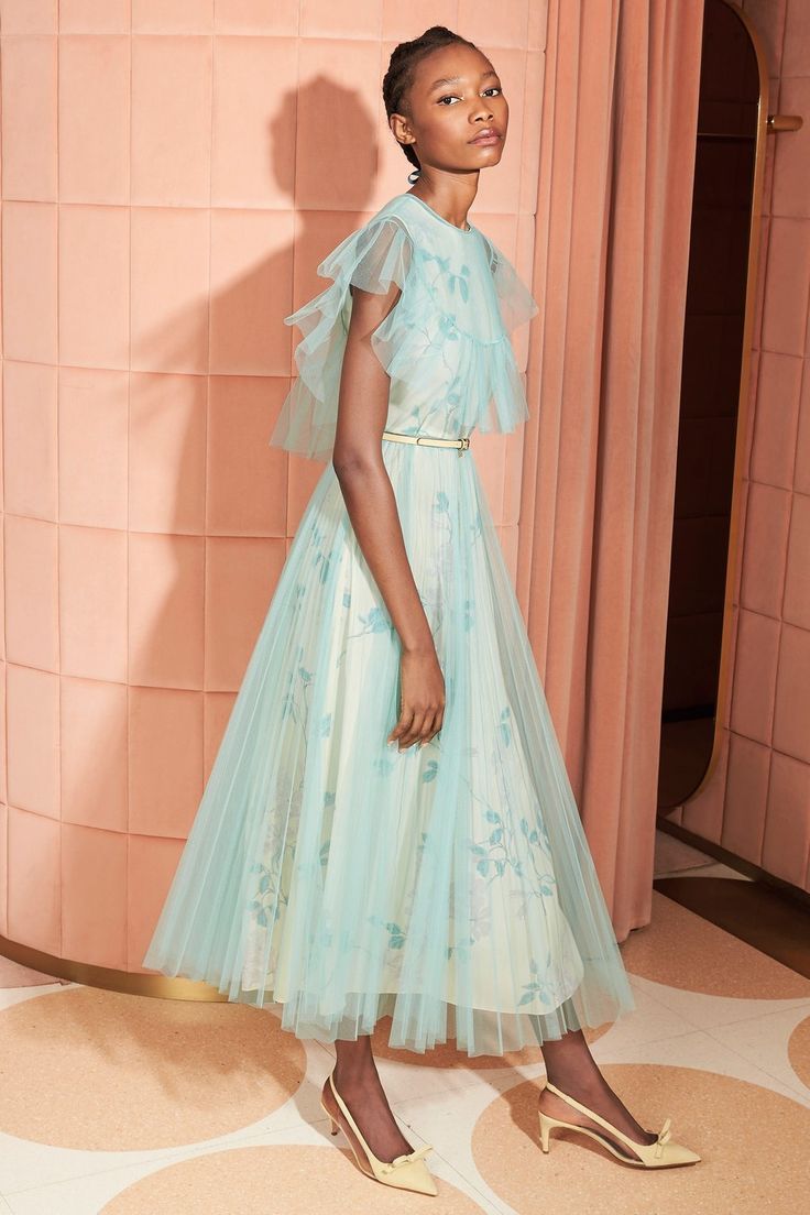 a young woman poses in a light blue pleated tulle midi dress with ruffled sleeves and a floral print. she is wearing pale yellow heels and is standing against a peachcolored wall with vertical paneling. the dress has a fitted bodice and a flowing skirt. the overall aesthetic is elegant, feminine, and stylish. the image is welllit and the model's pose is graceful.