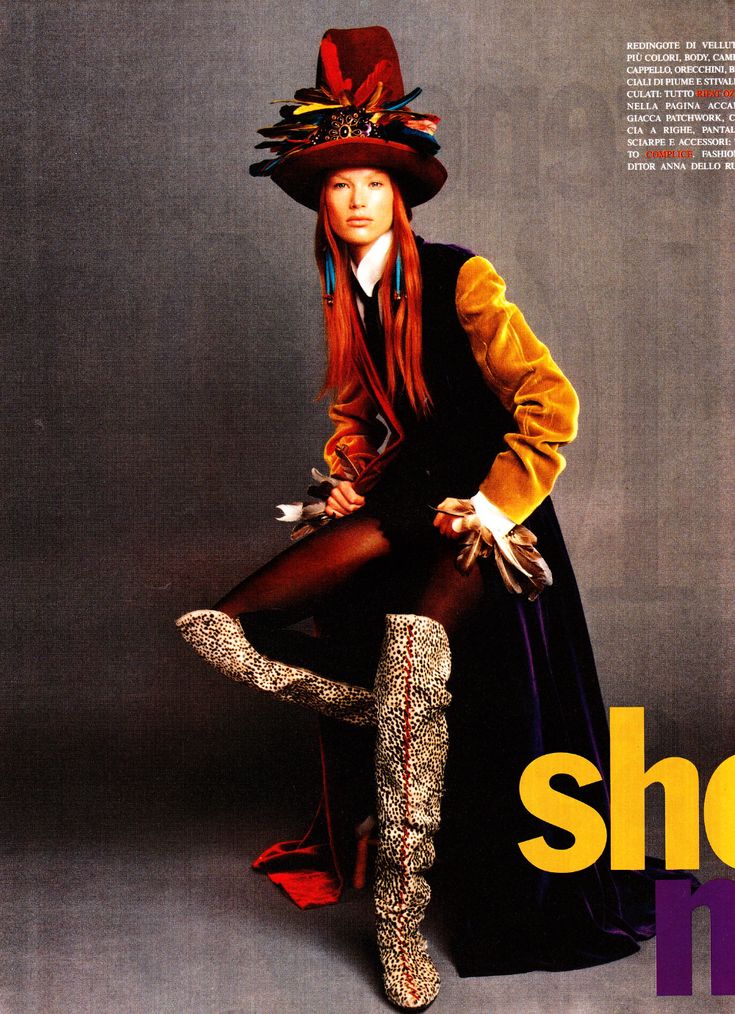 a fashion photoshoot of a model wearing a hat decorated with feathers, a dress with a yellow and black jacket, animalprint boots, and holding an object with feathers in her hand. the photo has a vintage magazine style and is against a grey background. some italian text is visible in the upper right corner.
