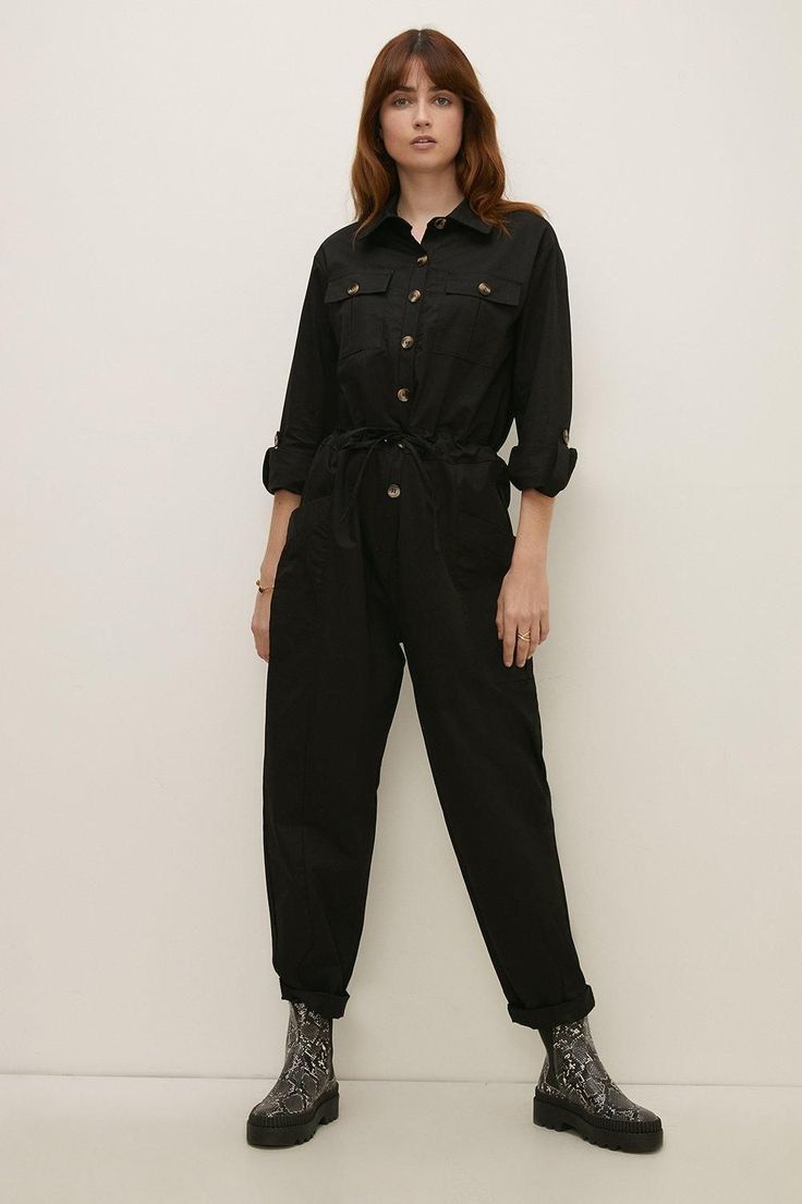 a young woman stands in front of a white background wearing a black longsleeved jumpsuit with buttons.  she has her arms at her sides and her hair is down.  she is wearing snake skin ankle boots.  she has a neutral expression on her face.