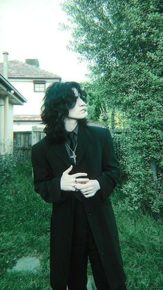 a woman with long black hair is standing in a dark suit and coat looking away. her suit has a white shirt and black tie. she is standing outside in front of a green background of trees and foliage.