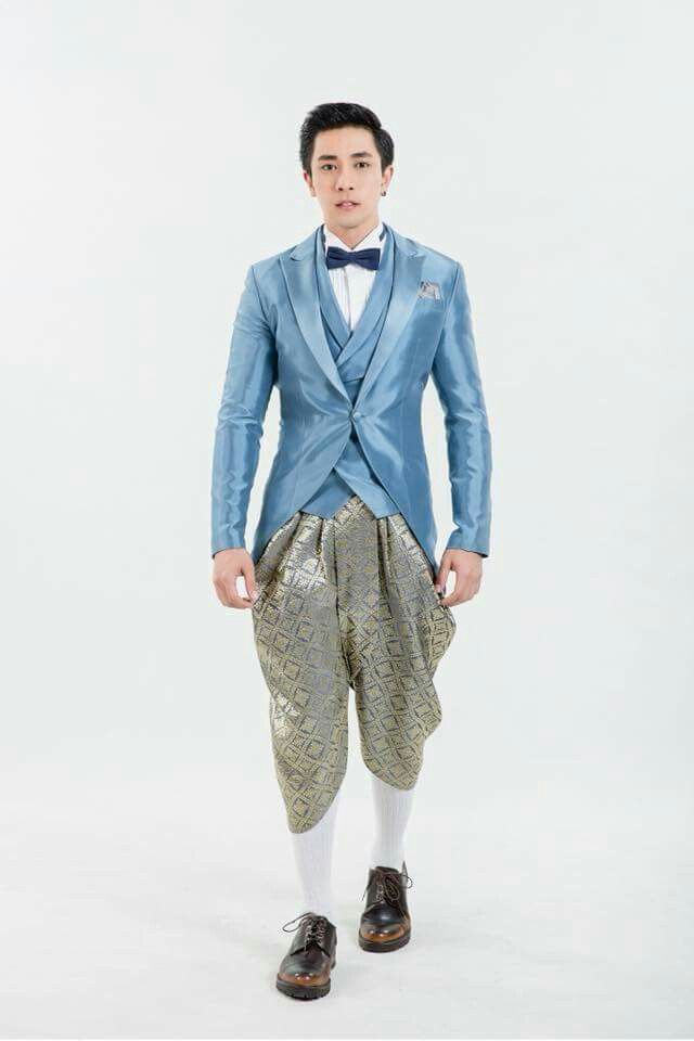 a young man in a blue suit with a bow tie and a white shirt is standing and looking at the camera. he is wearing traditional thai pants that are gold and blue and brown shoes. the background is white.