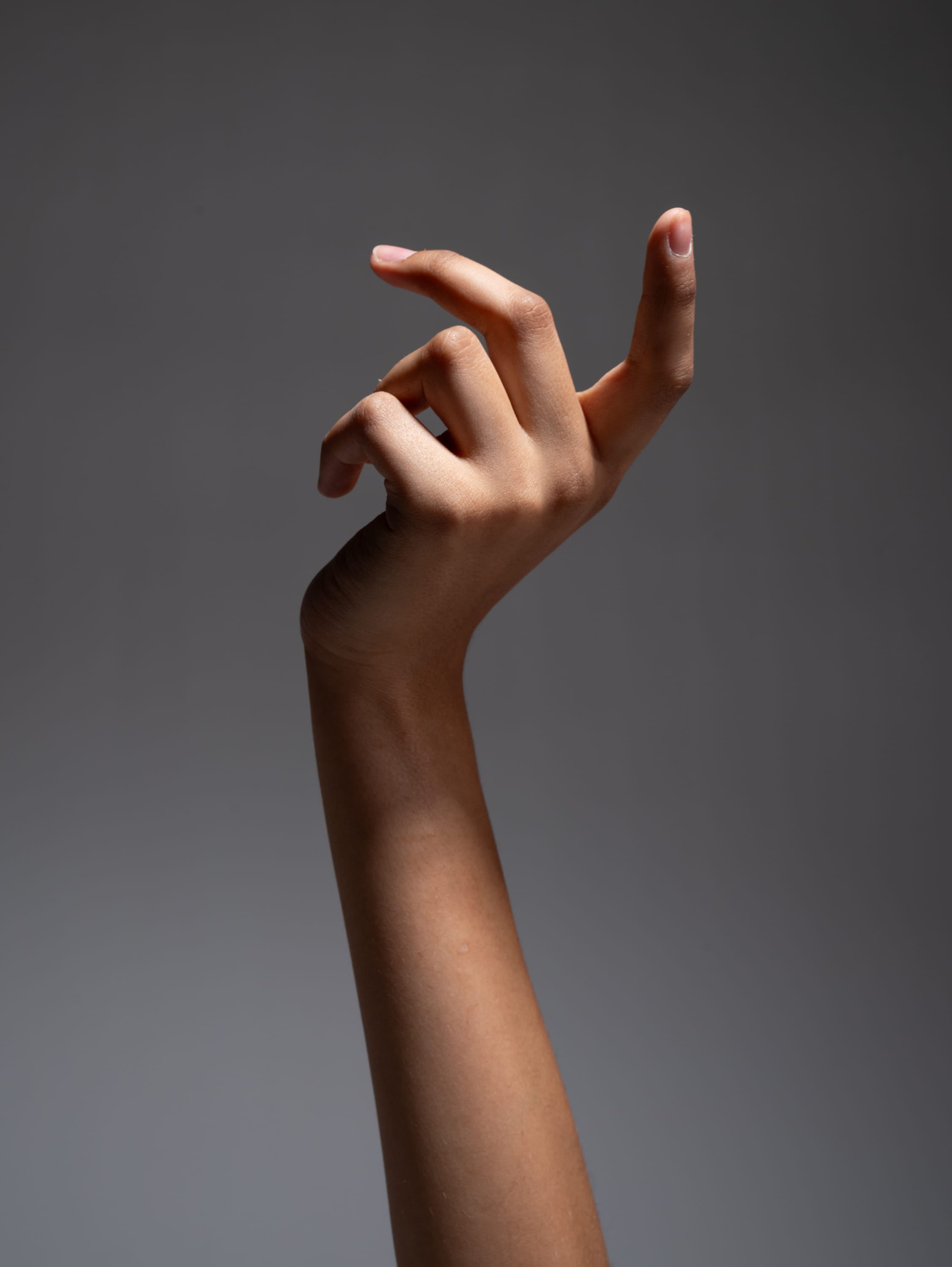 a fairskinned hand is reaching upward with the index and middle fingers slightly curled. the hand and arm are illuminated by a studio light against a gray background.