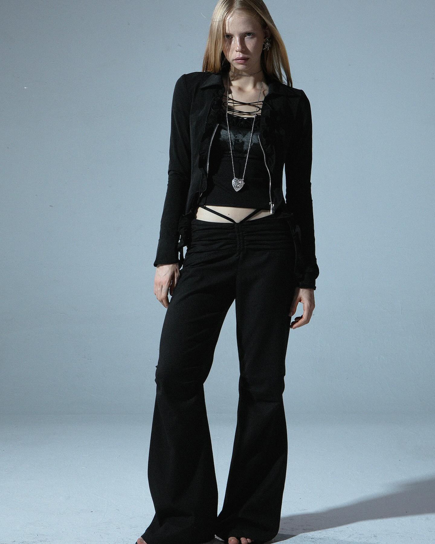 a fullbody shot of a blonde woman posing in a studio. she is wearing a black cropped jacket, a black laceup top, and black flared pants. she's accessorized with a silver heart necklace and earrings.  her hair is long and straight. the overall style is dark, alternative, and slightly gothic. the background is a simple, light gray.