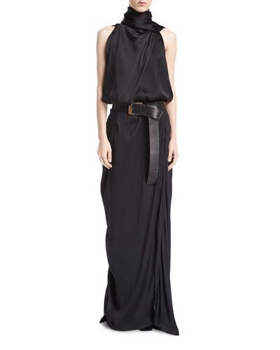 a woman in a black long, sleeveless, satin dress with a high neck and a black belt stands against a white backdrop. she is posing for a photo shoot.