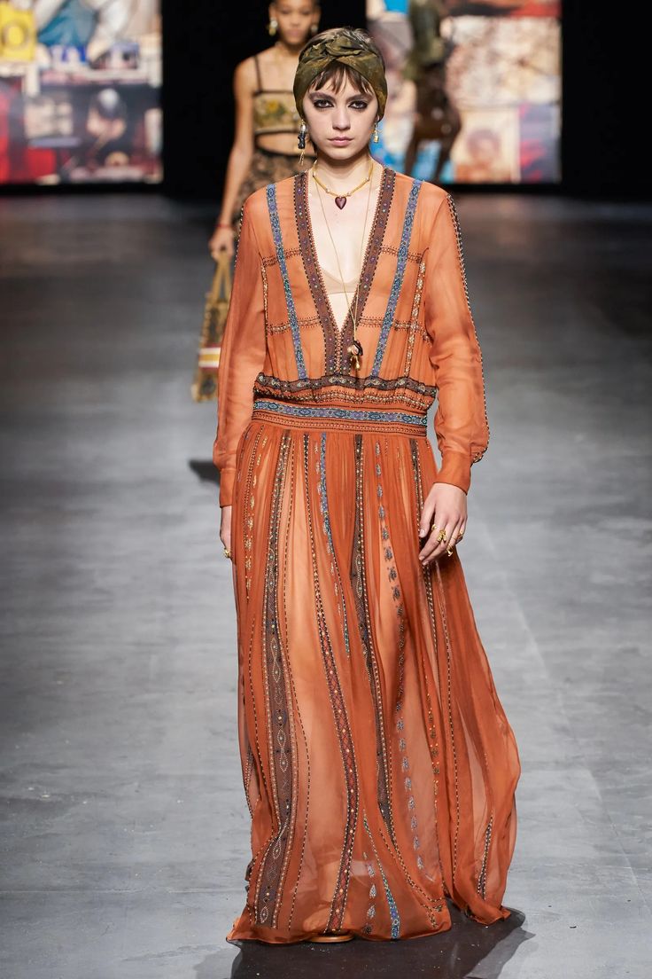 a model walks the runway in a long, flowing orange dress with intricate embroidery and brown embellishments. she is wearing a gold necklace and earrings, a head scarf,  and has her hair pulled back. the dress is semisheer, revealing a hint of the model's skin, and has a  flowy, bohemian vibe. the model is confident and stylish in her pose, showcasing the exquisite details of the dress and the designer's vision.  the runway is a blur of motion, highlighting the dress's graceful movement.