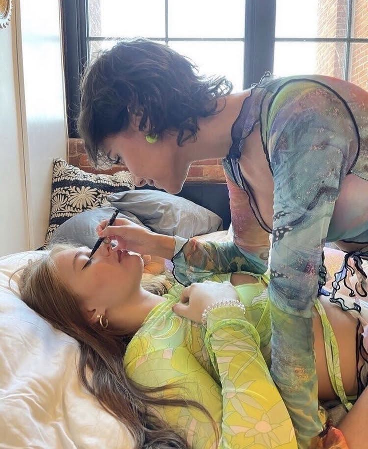 two women are laying on a bed with a white duvet. one woman is applying eyeliner to the other woman's eye. the woman who is applying the makeup is wearing a colorful, mesh shirt, and the other woman is wearing a green, mesh shirt.  the woman applying the makeup has short, dark, curly hair and is smiling while she looks at her friend. the woman having makeup applied has long, straight, dark hair and is looking away from the camera. there is a patterned pillow behind them.  the image is taken from the side, giving the viewer a clear view of the woman applying makeup.