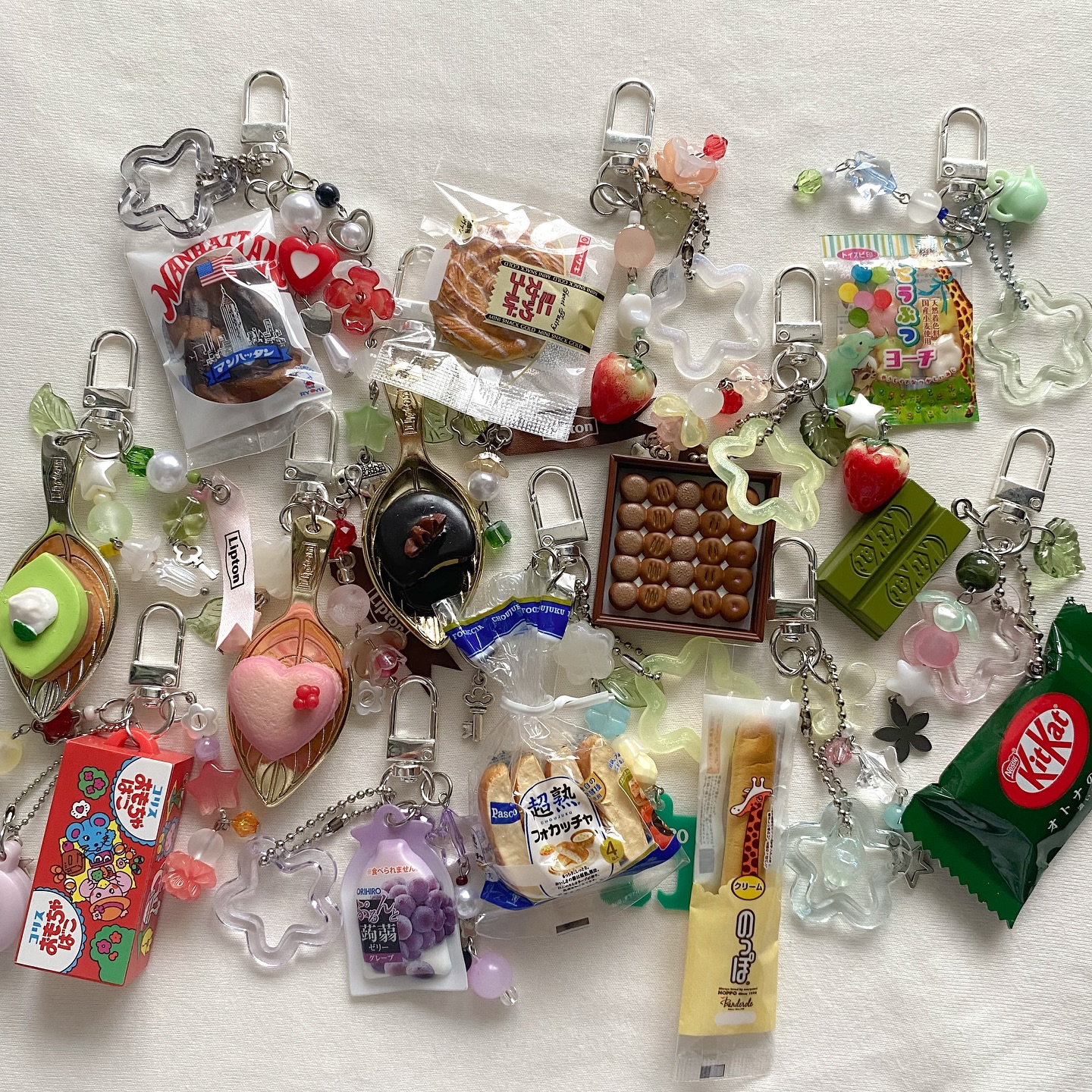 the image displays a collection of keychains featuring miniature food items, candies, and snacks. there are keychains with miniature packages of lipton tea, grapes, kiwi slices, kit kat bars, assorted candies, heartshaped objects, flowershaped ornaments, and various types of bread and baked goods. other keychains show miniature chocolates, donuts, starshaped decorations, packaging with cartoon characters, a giraffe illustration, and a package of peach focaccia. the arrangement of the keychains is scattered across a white background.