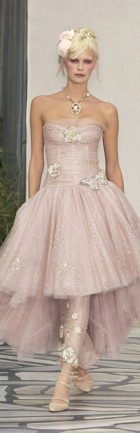 a fashion model walks down the runway wearing a stunning, intricately embellished, pink tulle gown. the gown features floral accents, sequins, and a layered skirt with a highlow hemline. the model's hair is styled in a loose updo and adorned with delicate flowers, and she is accessorized with a pearl necklace, silver earrings, and sparkling makeup. the model exudes elegance and femininity, capturing the essence of a glamorous evening look.
