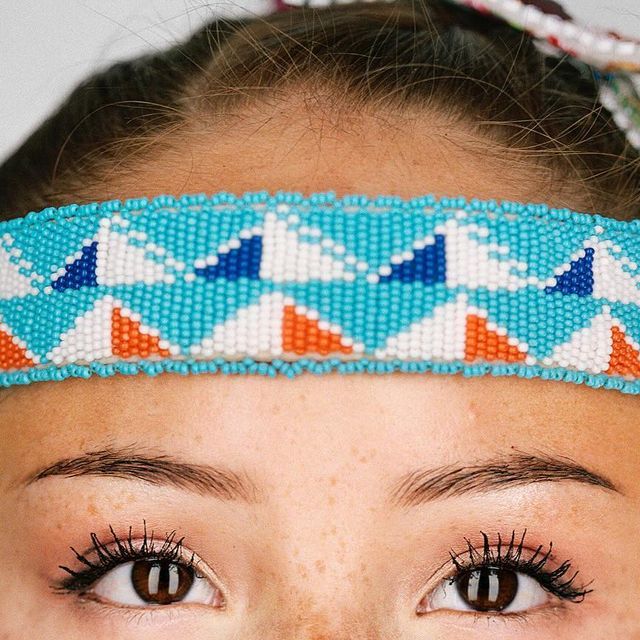 a closeup shot of a young woman's face, the focus is on her eyes and the vibrant beaded headband across her forehead. the headband features a geometric pattern primarily in turquoise, orange, white, and dark blue triangles. the woman's skin is lighttoned with freckles visible.  her hair is pulled back, and her makeup is subtle. the overall impression is one of beauty and cultural expression.  the style of the headband suggests influences from native american art and beadwork.