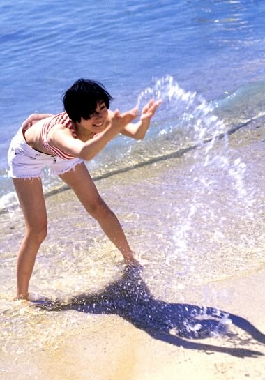 a woman with short black hair is playing in the shallow water at the edge of a beach. she is wearing a red and white striped top and white shorts. she is bending over and splashing water with her hands. the sunlight creates a dark shadow of her on the wet sand. the ocean is visible in the background.