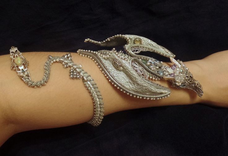the image shows an ornate, silvercolored arm jewelry piece on a person's arm. it consists of a braceletlike element that wraps around the arm in a snakelike pattern, adorned with beads and a gemstone. the design extends into a more elaborate, clawlike structure that covers the wrist and part of the hand, featuring intricate metalwork, pearls, and potentially other gemstones. the overall impression is one of fantasy, gothic, or perhaps tribalinspired fashion.