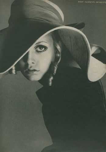 a black and white portrait of a woman wearing a widebrimmed hat, her face partially obscured by the hat's shadow. she is looking directly at the viewer, her expression is both alluring and mysterious. the image is likely from a fashion magazine and showcases the popular style of the 1960s and 1970s.