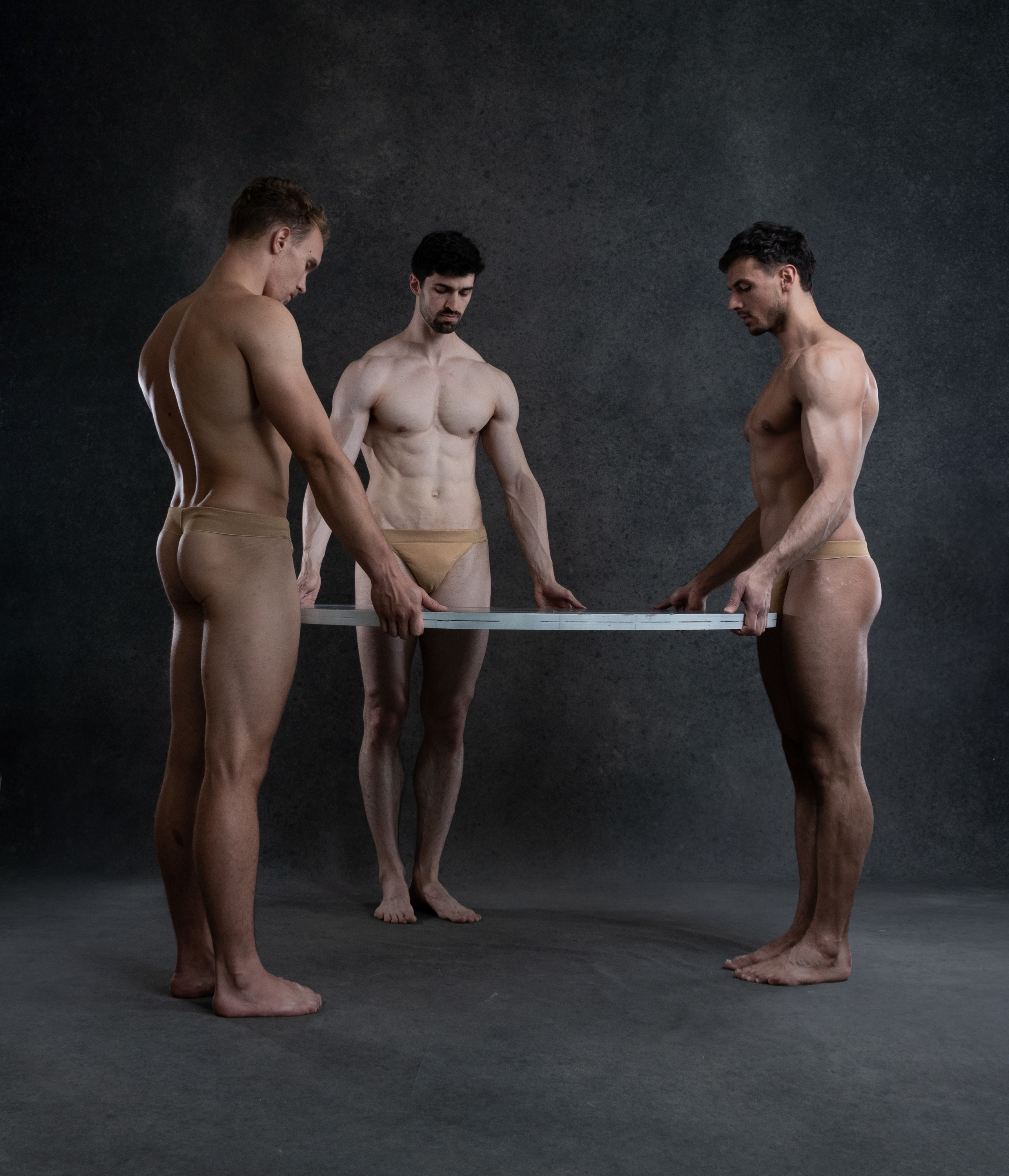 three muscular men in nude color underwear pose in a studio setting with dark gray background. the men are holding a long rectangular object (light or frame) in front of them at hip level, with their hands on either side of it.  the man on the left is looking down at the object, while the other two men are looking straight ahead.  they are lit by studio lighting.