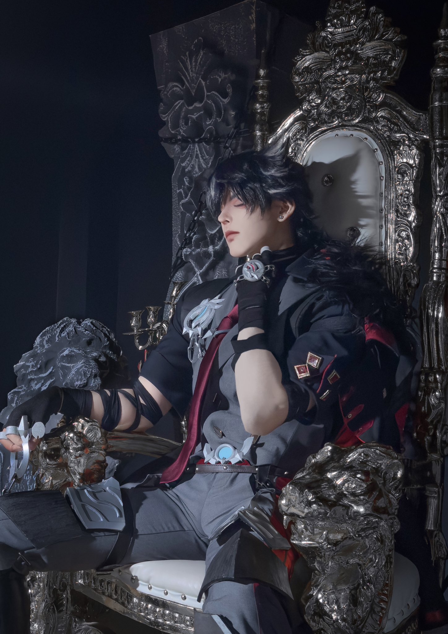 this image shows a young man cosplaying as a dark and mysterious anime character. he is sitting on a large, ornate throne and holding a sword in his hand. he is dressed in dark clothing with a silver belt and gloves. the image is dark and dramatic, with a sense of intensity about it.