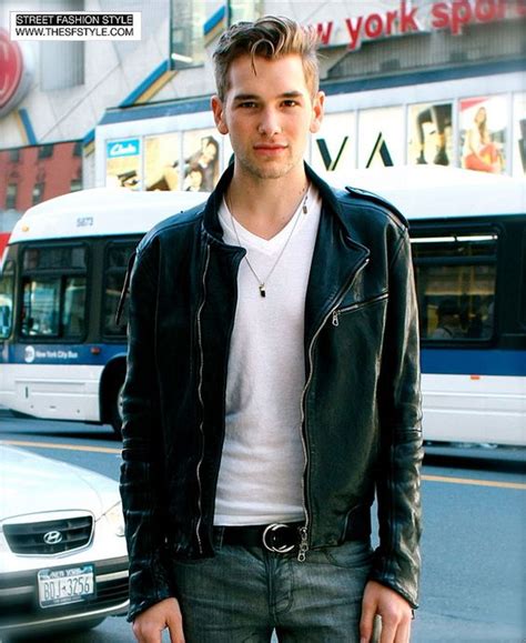 a young man with styled hair stands on a city street, posing for the camera. he's wearing a black leather jacket over a white tshirt, paired with dark wash jeans and a silver belt. he's positioned slightly offcenter to the left, with a city bus and urban buildings visible in the background. the overall image suggests a street style fashion aesthetic.