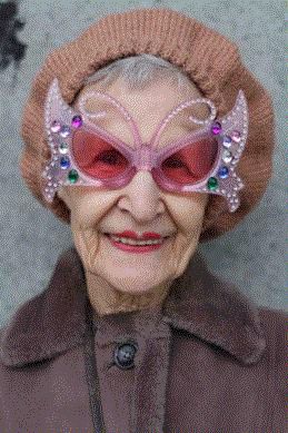 an elderly woman wearing a brown knitted hat and a brown coat is smiling at the camera. she is wearing butterflyshaped glasses with pink lenses and sequins around the edges.