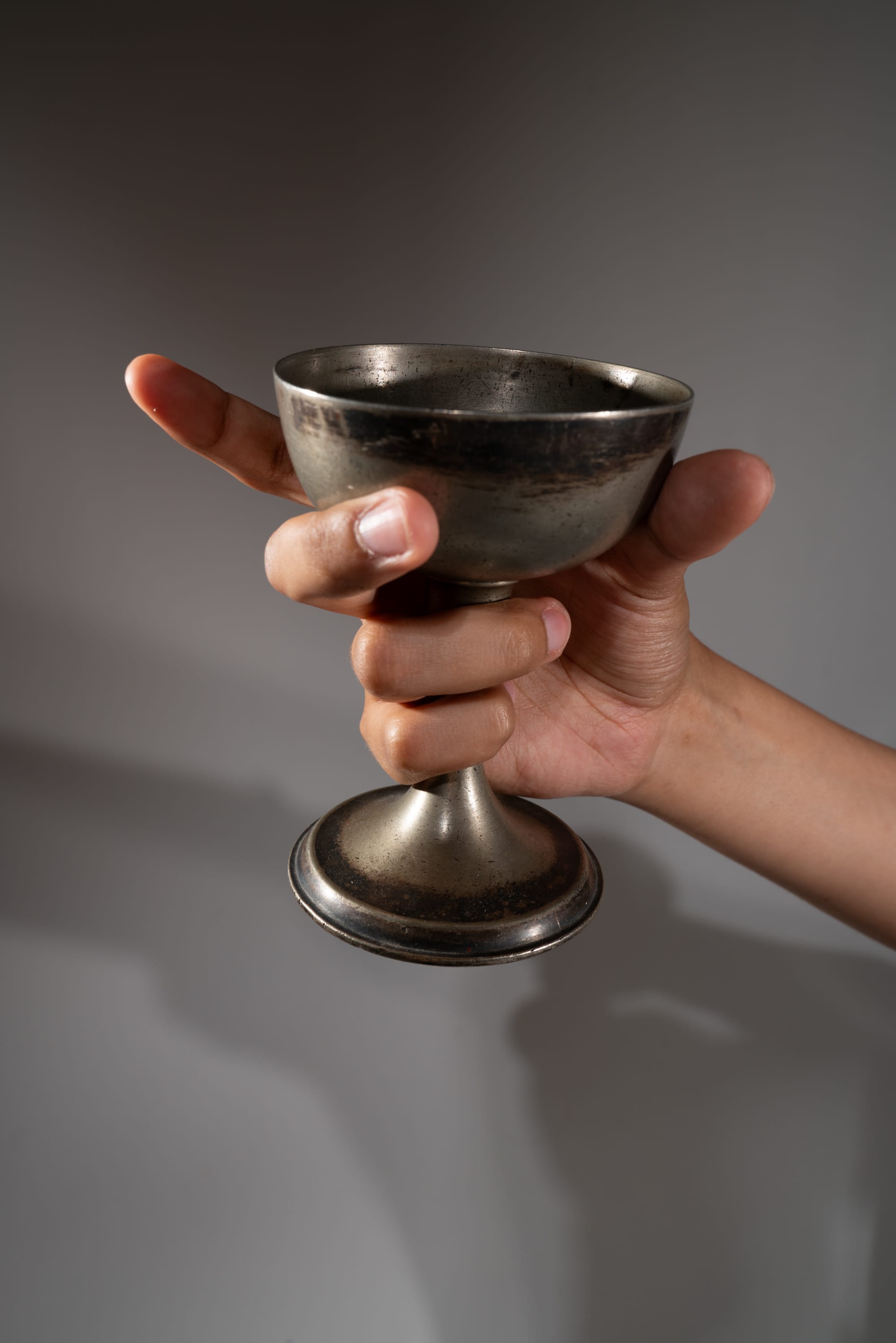 a hand holds up a tarnished silver chalice against a gray background. the hand is slightly out of focus. the chalice is detailed, with a wide bowl, a stem, and a round base.