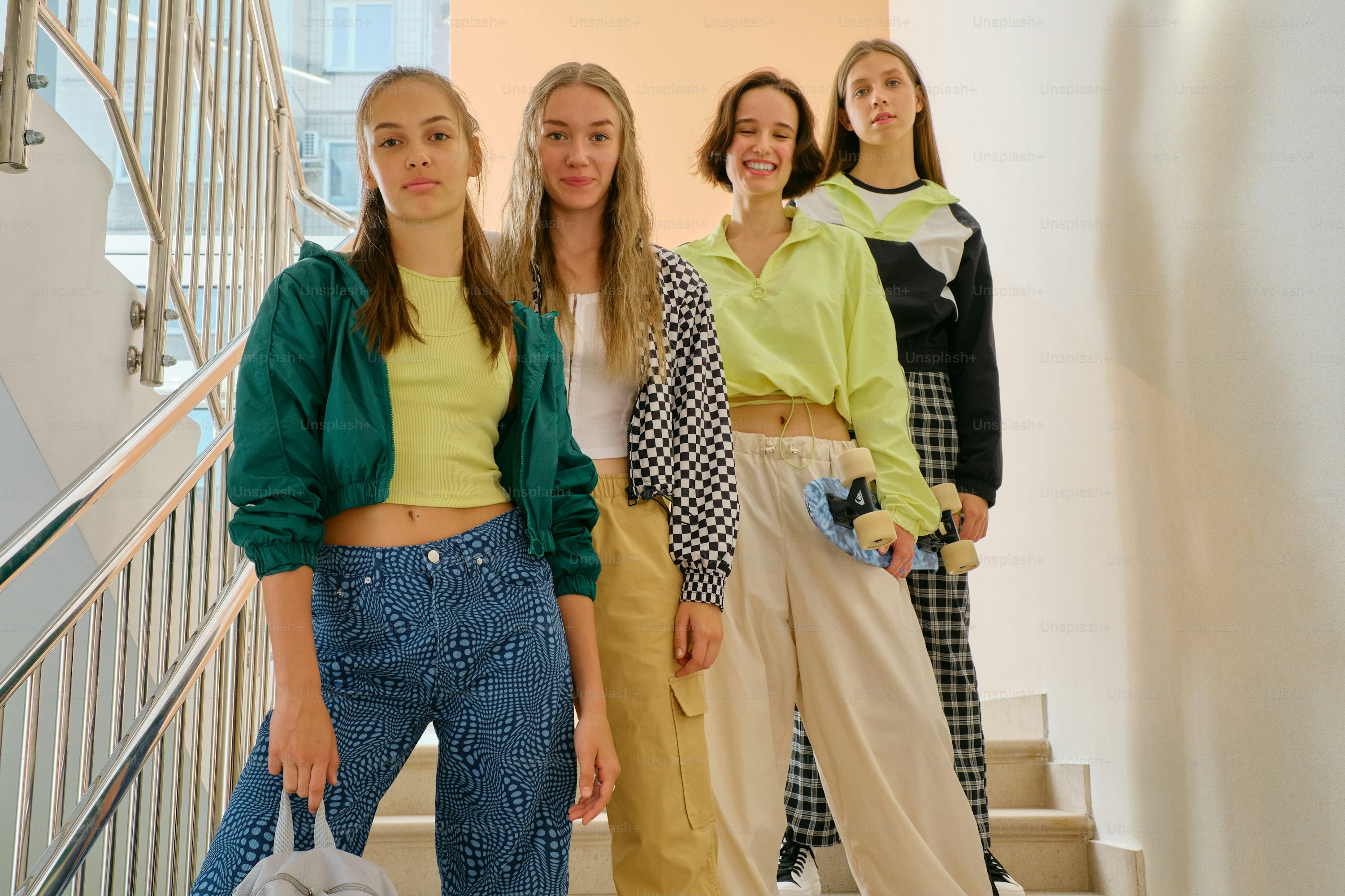 a group of four teenage girls stand on a staircase, smiling at the camera. they are dressed in stylish, casual clothing, including colorful pants, jackets, and crop tops.  some of the girls are carrying skateboards. the setting appears to be indoors, possibly in a school hallway. the overall mood is one of friendship and youthful energy.