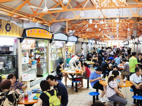 the image depicts a bustling and vibrant hawker center in singapore. numerous food stalls line the walkways, each offering a variety of dishes.  crowds of people are seated at tables, enjoying their meals.  the atmosphere is lively and energetic, showcasing the social and cultural aspect of eating at a hawker center. the architecture of the center is visible, featuring an orangecolored metal roof structure. the overall scene is one of a typical, busy, and lively day in a singaporean food court.