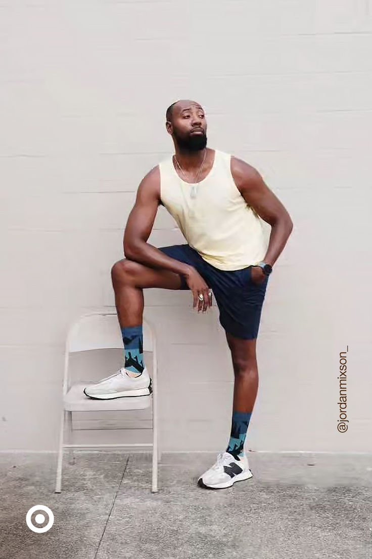 the image shows a stylish black man posing outdoors against a lightcolored wall. he's wearing a yellow tank top, dark navy shorts, white sneakers, and eyecatching patterned socks. his pose is relaxed and confident, with one leg resting on a white chair. the overall style is casual and summery. the photo is welllit and captures a candid, fashionable vibe. the instagram handle "@jordannmixson_" is visible in the image's bottom right corner.