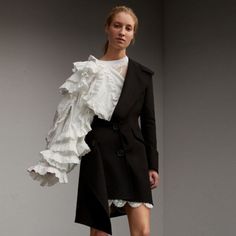 a woman is wearing a white ruffled shirt with a black blazer. the blazer has a white lace trim and a black button on the bottom. the woman is standing in front of a gray background and looking at the camera. she has long blonde hair and her makeup is minimal. the image is taken from a slightly high angle.