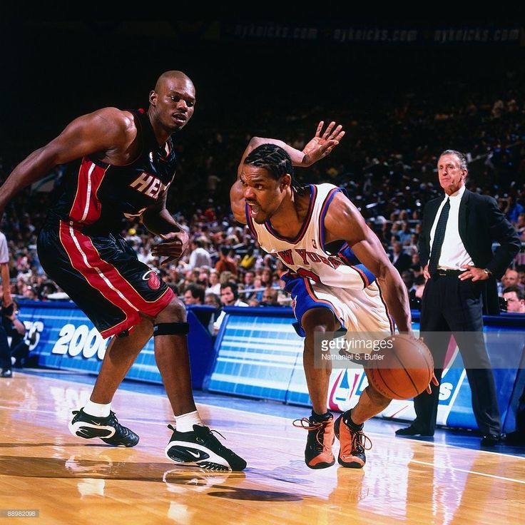 the image is an action shot from an nba basketball game.  it features allan houston of the new york knicks, dribbling the ball intently while being closely guarded by p.j. brown of the miami heat. the background shows a blurred crowd and the coach of the knicks watching the play.  the focus is sharply on the two players, highlighting their intense concentration and the physicality of the game. the lighting and photography style suggest it's a professional sports photograph, likely from the 1990s given the players' attire and court signage.