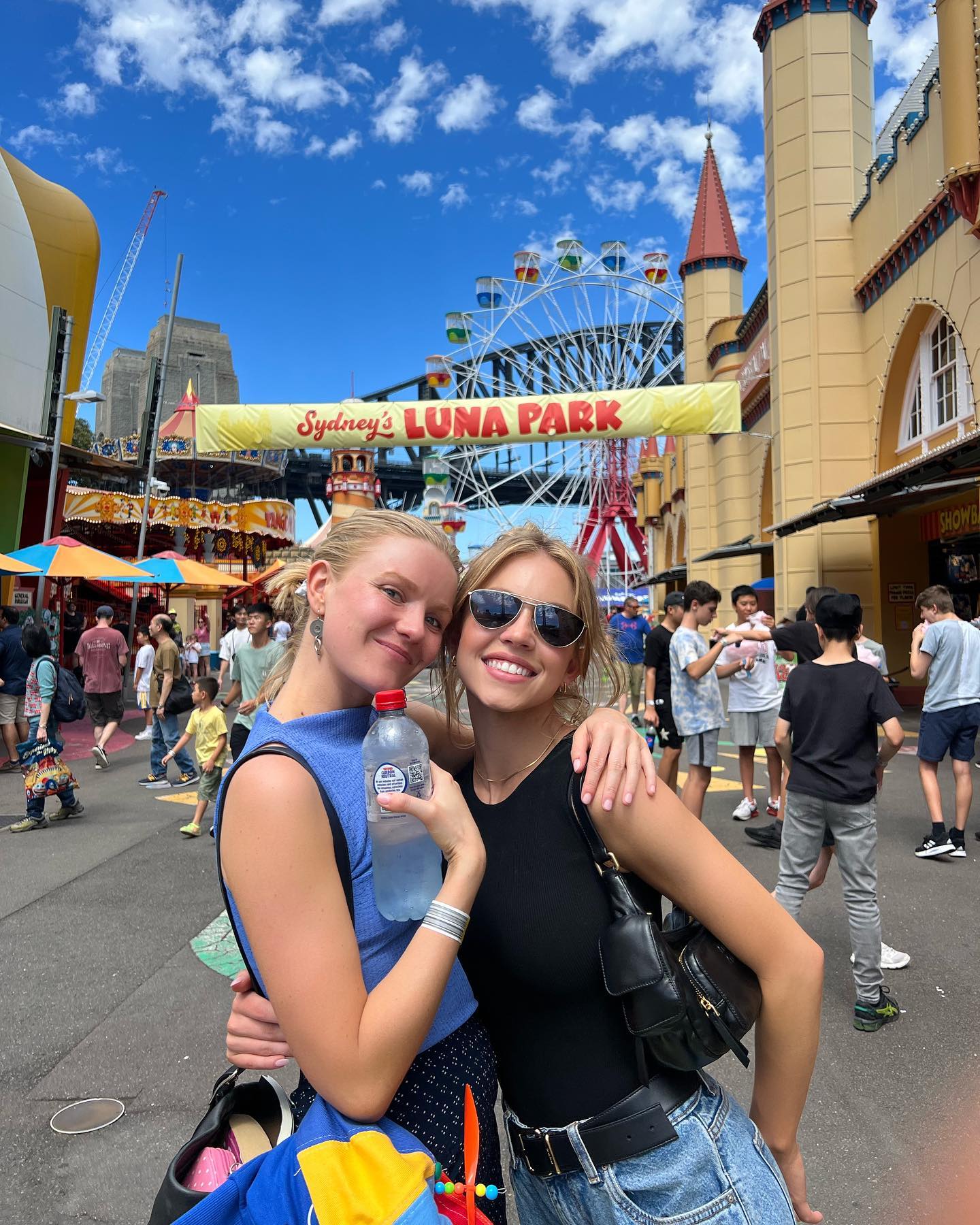 two teenage girls are smiling happily for a picture at sydney luna park on a sunny day. the iconic ferris wheel and harbor bridge are visible in the background, along with other amusement park rides and attractions.  the girls are dressed casually and are hugging each other.  a bustling crowd of people can be seen in the background, enjoying the amusement park. one girl is holding a bottle of water. the overall atmosphere is vibrant and cheerful.