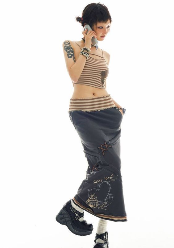 a fullbody shot of a young woman posing against a white background. she is wearing a brown striped tank top, a long gray maxi skirt with brown details, and black platform boots. she is holding a cell phone to her ear and has tattoos on her arms. her hair is styled in a short, layered cut.
