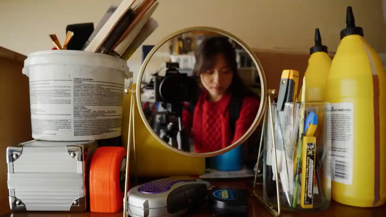 a woman takes a selfie in a workshop setting, her reflection captured in a round mirror. she wears a red sweater and has dark hair. surrounding her are various tools and art supplies, including paint cans, brushes, a measuring tape, and a level. the warm, natural light suggests a daytime setting.  the overall impression is one of creativity and a handson approach to making.