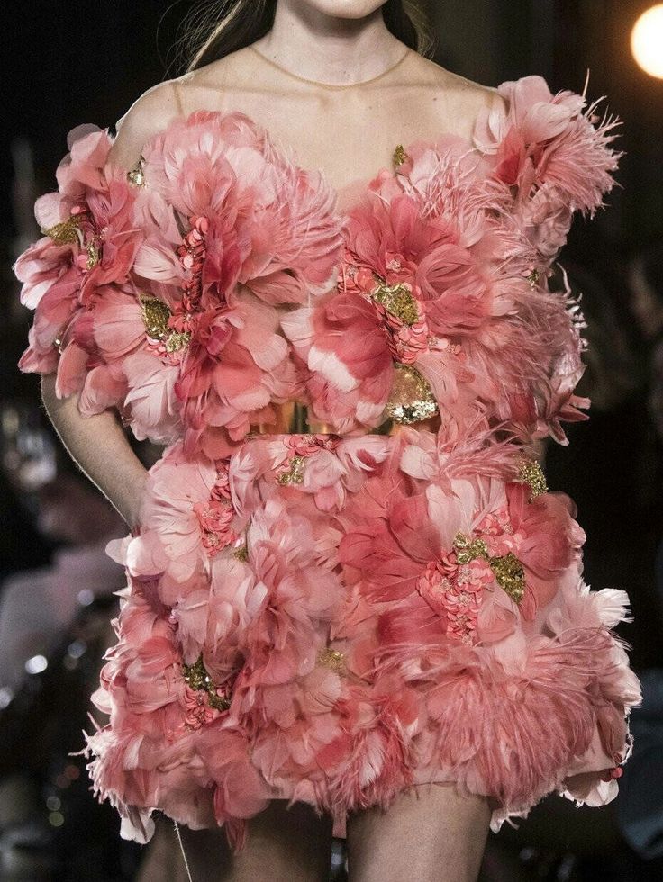 a closeup shot of a model wearing a pink dress made of feathers and flowers, walking the runway during a high fashion show. the dress is extremely detailed and elaborate, with delicate feathers and intricate floral embellishments. the model's hair is pulled back, and she is wearing minimal makeup, allowing the dress to take center stage. the overall aesthetic is romantic and ethereal.