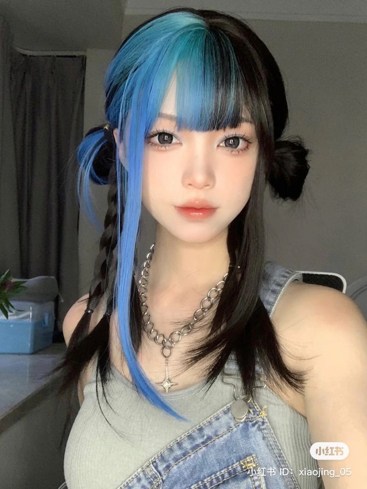 a young asian woman poses for a selfie, showcasing her trendy splitdyed hair. her hair is styled in halfup space buns with braided sides. the left side of her hair is dyed a vibrant blue and teal, while the right side is her natural black.  she wears minimal makeup and a simple silver chain necklace, her denim overalls completing the casual yet stylish look. the photo is taken indoors with natural lighting.