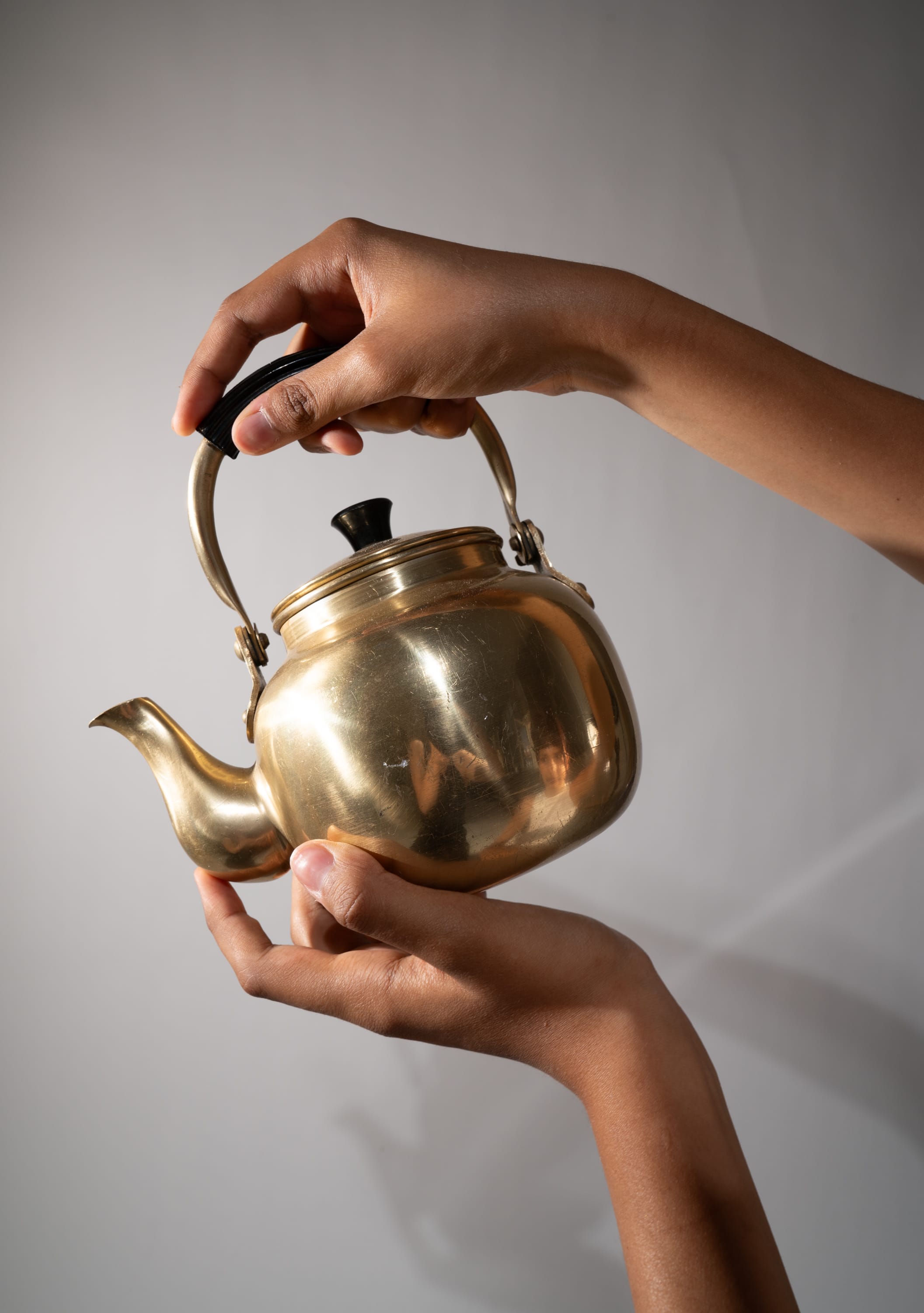 a closeup shot of a person holding a vintage brass tea kettle against a white background. the kettle has a black handle and knob, and the brass is polished to a shine. the person is holding the kettle by the handle and the lid, and their hands are visible in the foreground. the image has a warm and inviting feel, evoking a sense of nostalgia and tradition.