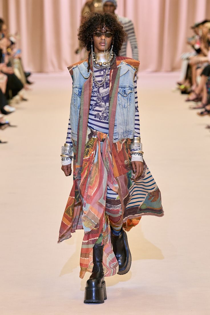a female model walks down a runway in a colorful and stylish outfit. she is wearing a denim jacket, a colorful skirt, and a pair of chunky black boots. she is also accessorized with sunglasses, a necklace, and bracelets.