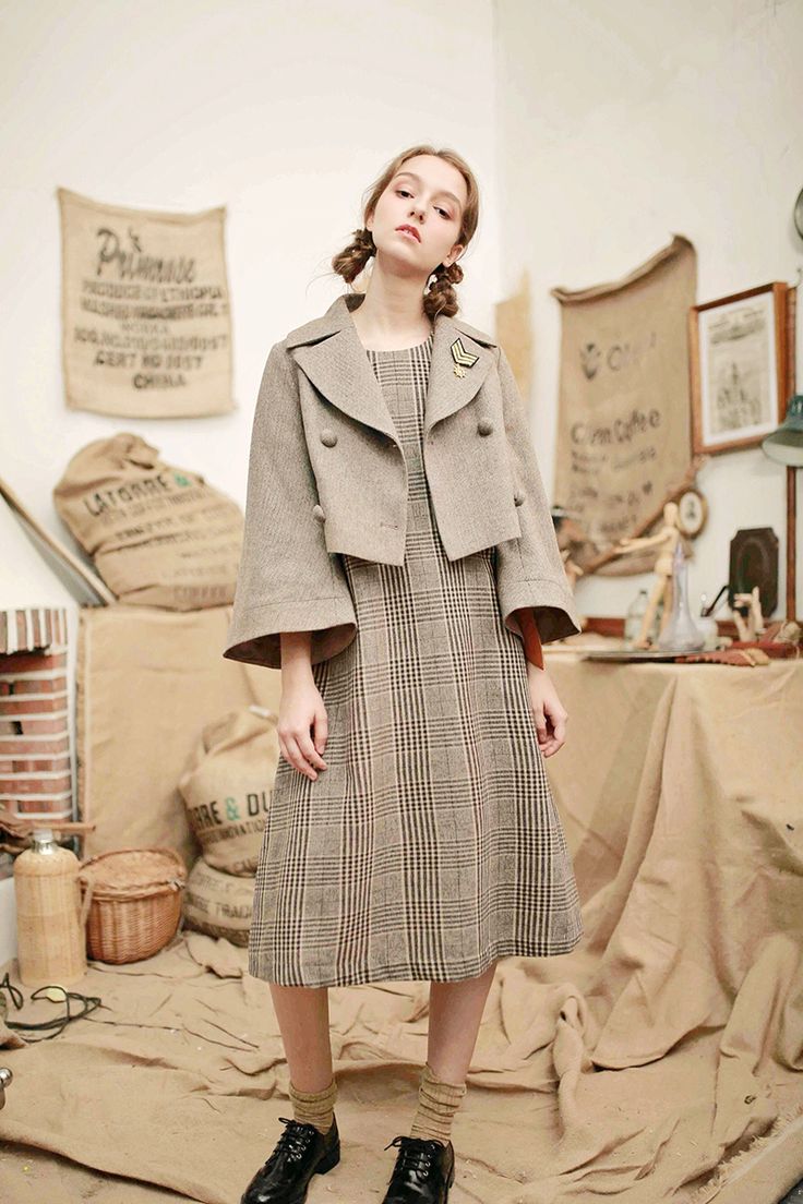 a fullbody shot of a young woman modeling a stylish outfit in a studio setting. she is wearing a beige cropped plaid coat over a matching midi dress with a classic checkered pattern.  her accessories include a small brooch on her jacket and dark brown oxford shoes with socks. the backdrop is rustic, featuring burlap sacks, wooden crates and other vintage props that create a retro, almost bohemian atmosphere. the overall style is reminiscent of vintage fashion.