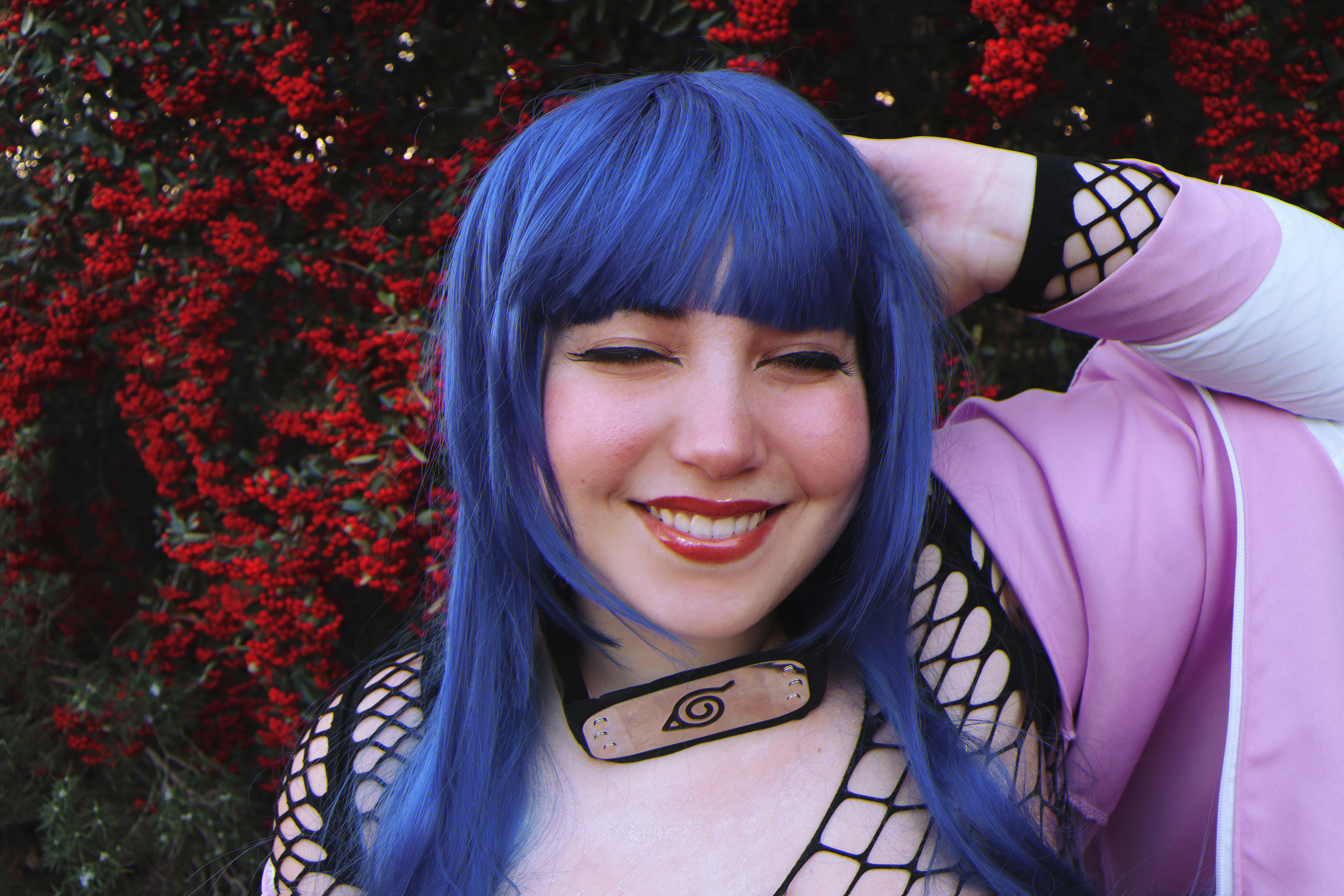 a woman dressed as hinata hyuga from the anime naruto is smiling at the camera. she is wearing a blue wig, forehead protector, and fishnet top. she has one hand on her head. in the background there is a bush with red berries.