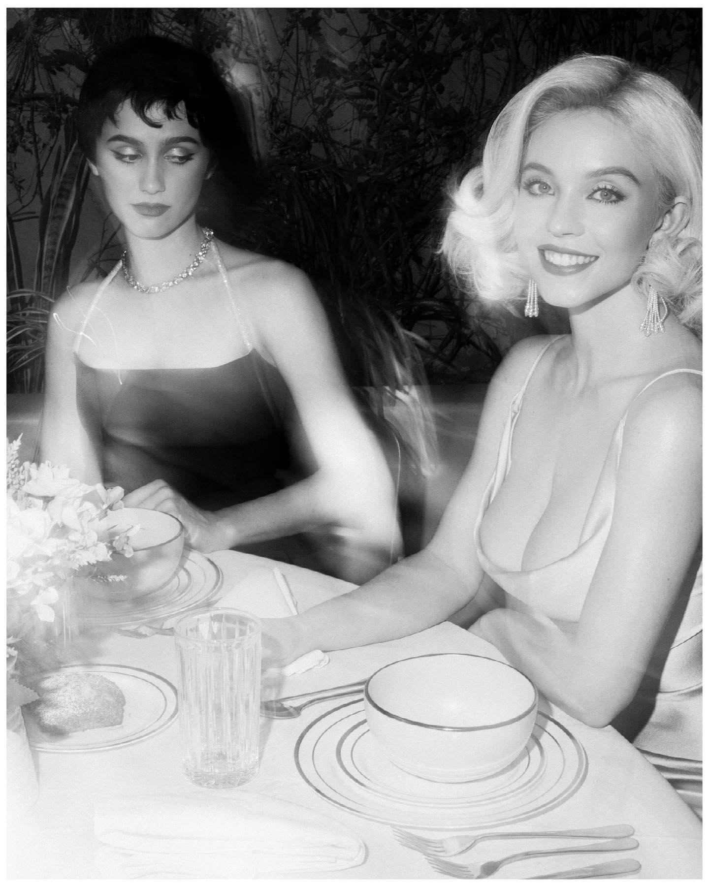 the black and white photograph shows two elegantly dressed women seated sidebyside at a table set for dinner.  the woman on the left has short dark hair and is wearing a darkcolored, possibly velvet, sleeveless dress with a halterneck style. she has a necklace and appears more reserved. the woman on the right has blonde hair styled in a voluminous, classic 1950sesque updo and is wearing a lightcolored, lowcut, sleeveless dress, which reveals some cleavage. she has dangling earrings and a more confident, outwardfacing demeanor. the setting appears upscale; there is fine tableware present  bowls, plates, and silverware  suggesting a formal occasion.  the background is blurred but indicates a lush, possibly outdoor setting. the overall mood is glamorous and vintage.  the image is styled in a way that resembles vintage hollywood photography.