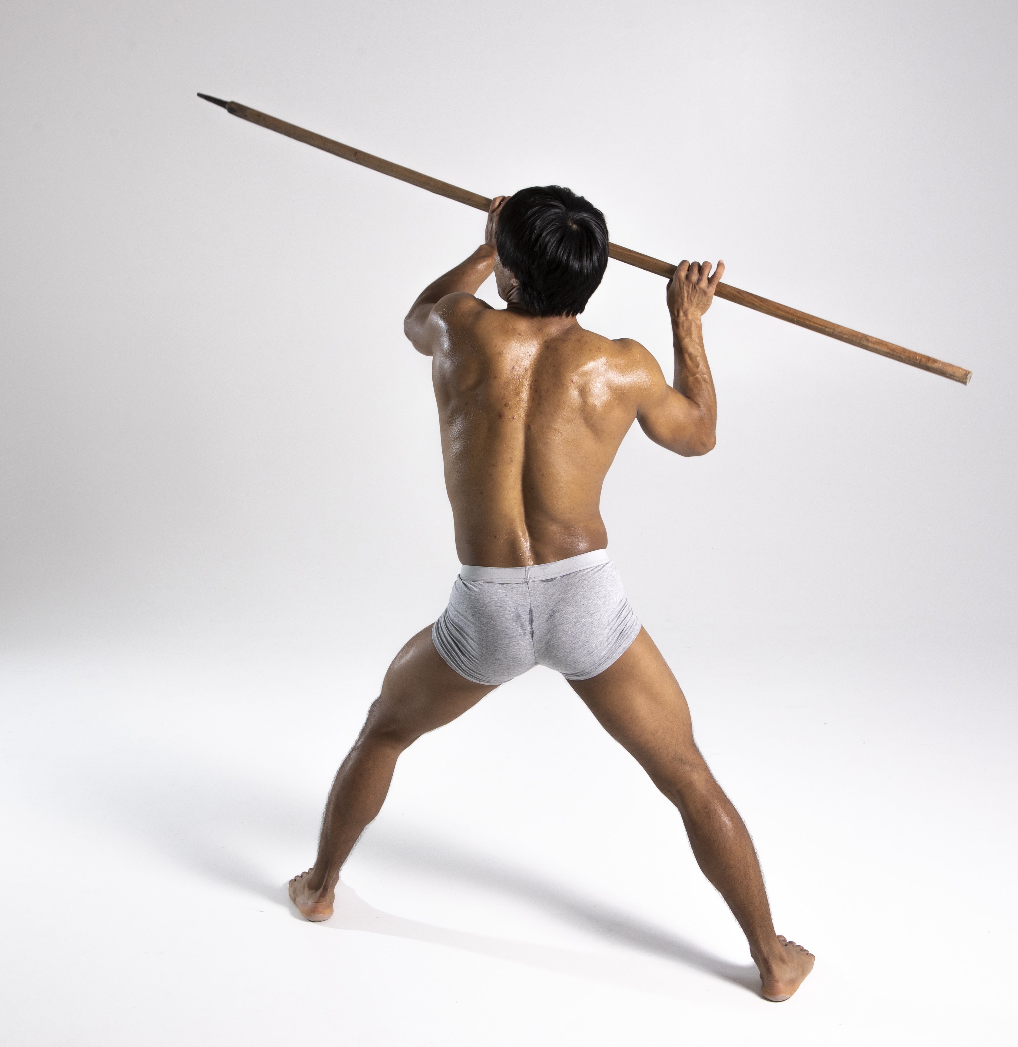 a shirtless man stands barefoot in a studio with a white background, holding a wooden spear overhead in a poised stance, revealing his muscular back and legs. he is only wearing grey underwear.
