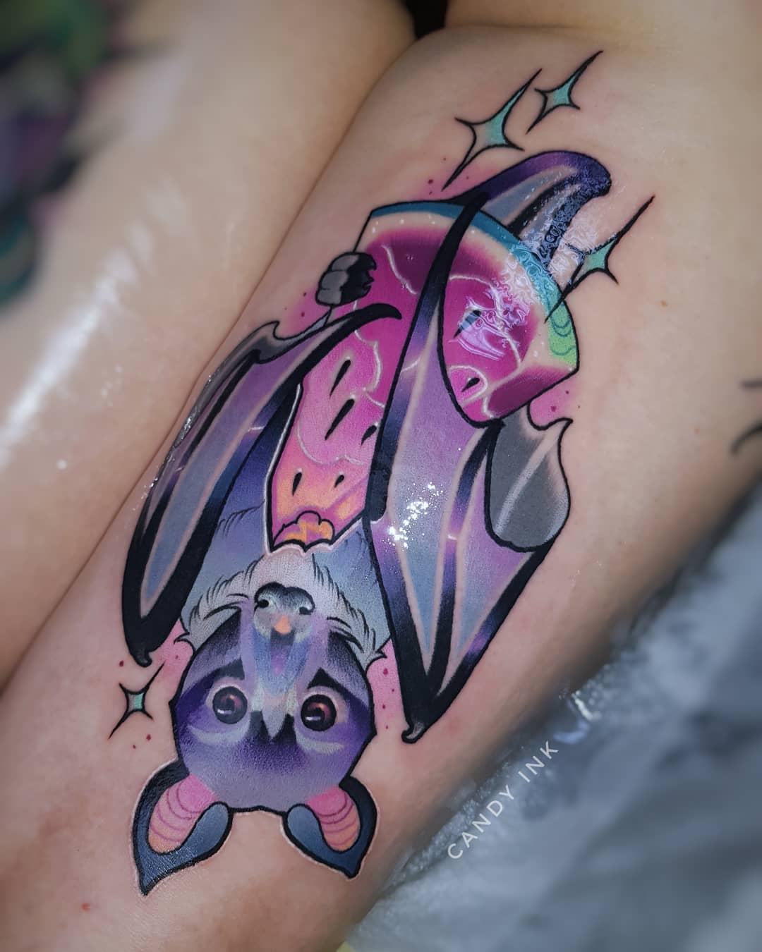 a person has gotten a neo traditional tattoo of a bat with its wings spread. the tattoo is on their upper thigh. the bat's wings are designed to look like slices of watermelon. the tattoo is colorful and the bat has large, cute eyes. the tattoo artist's signature, "candy ink", is visible in black ink on the bottom right of the tattoo.