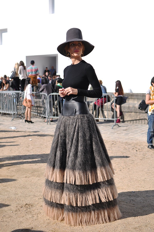 a stylish woman stands outdoors, dressed in a striking fashion ensemble. she wears a widebrimmed hat and a longsleeved black top, accentuated by a leather belt at the waist. her most distinctive piece is a multitiered skirt with a textured appearance. the scene is a public outdoor area with other people in the background.
